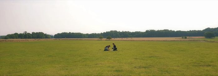 wide angle photo of two people kneeling in a grassy field, examining something on the ground
