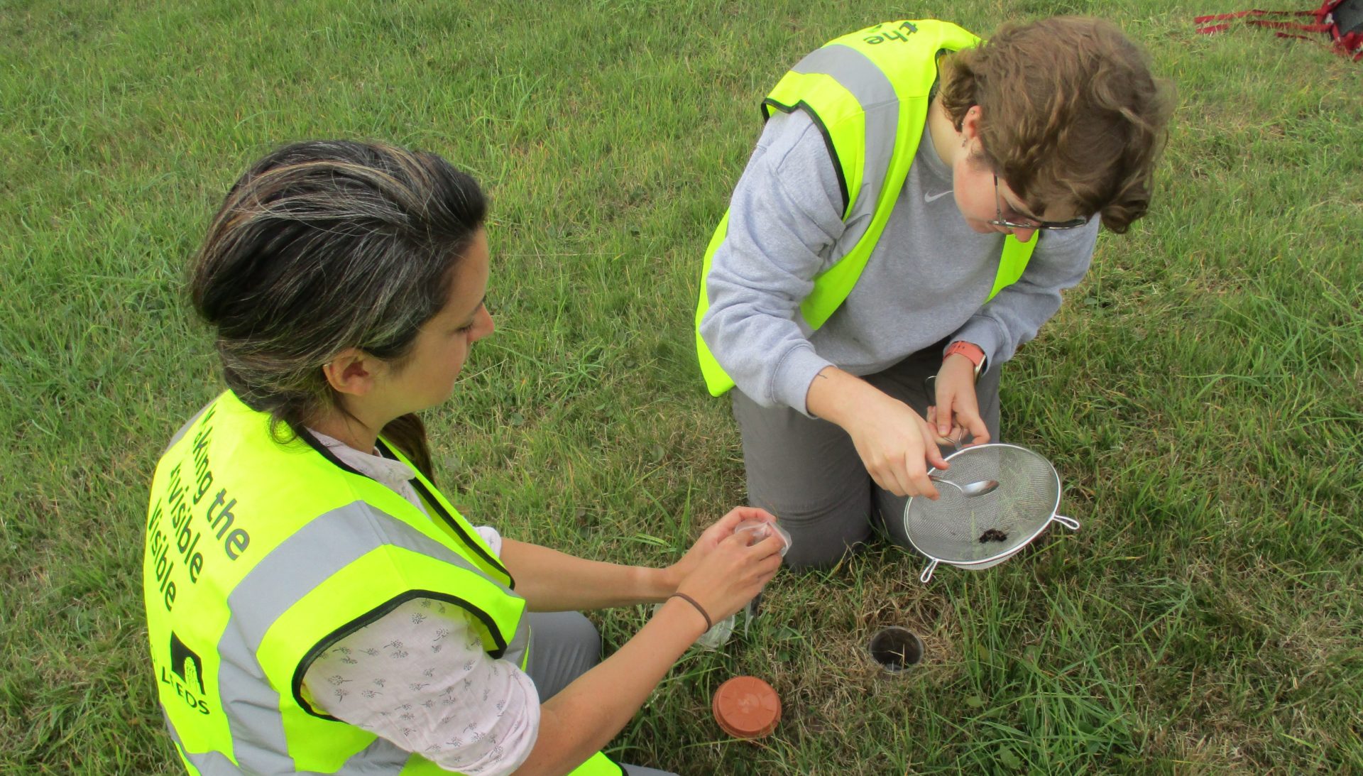 Two people in high visibility jackets looking at samples from a hole in the grassy ground in a sieve