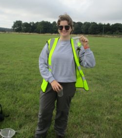 Alana in a high-visibility jacket holding up a sample bag containing vegetation